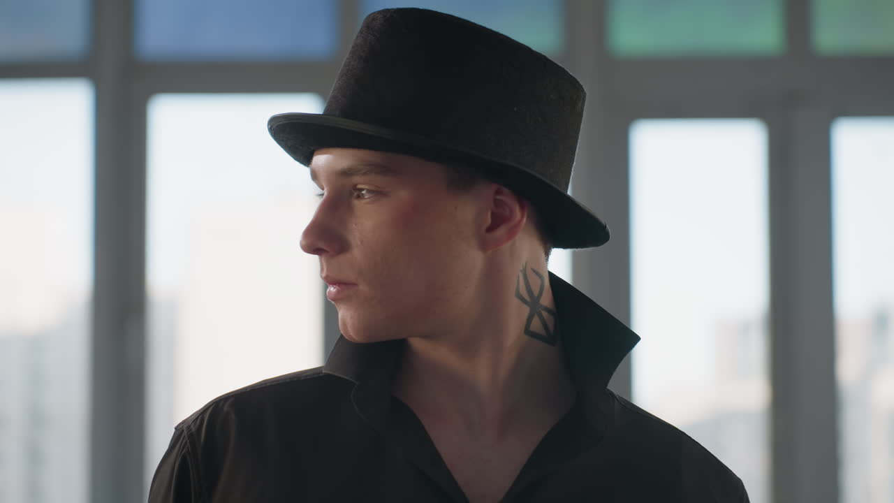 elegant young man in black shirt and hat tipping brim to display striking neck tattoo in front of loft window and mirror reflecting angular architecture moody natural light confident stylish gesture