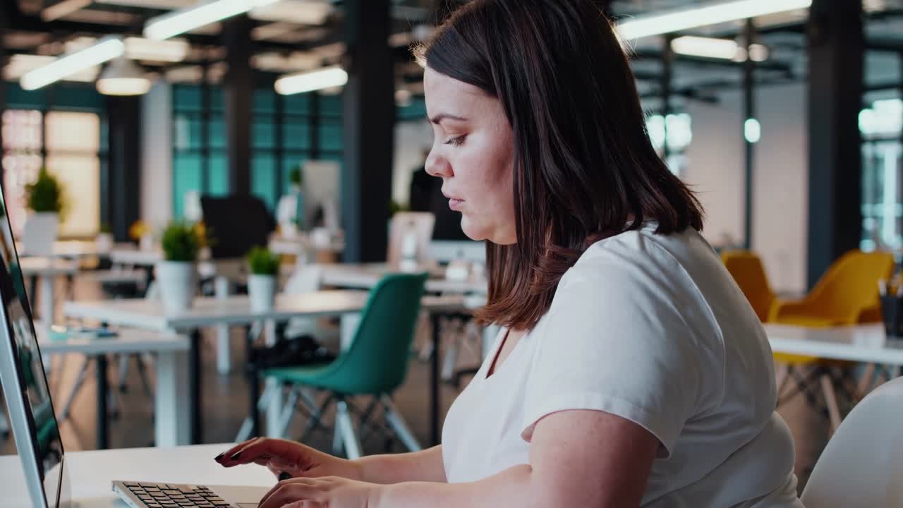 Side angle video of a woman focused on typing at a desk in a modern office