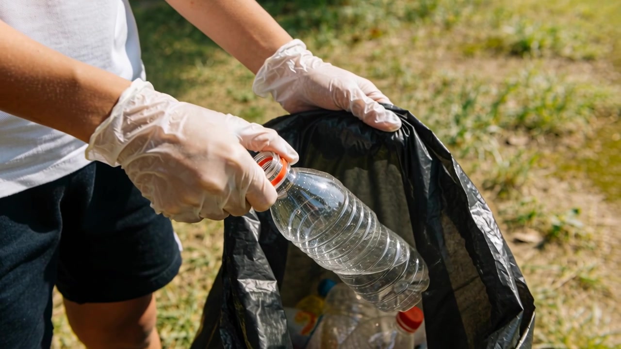 Picking up bottles, gloved white-shirt volunteer placing them into black bag at park for cleanup