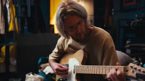 a blond man with long hair in glasses with a beard plays the electric guitar in a cozy room