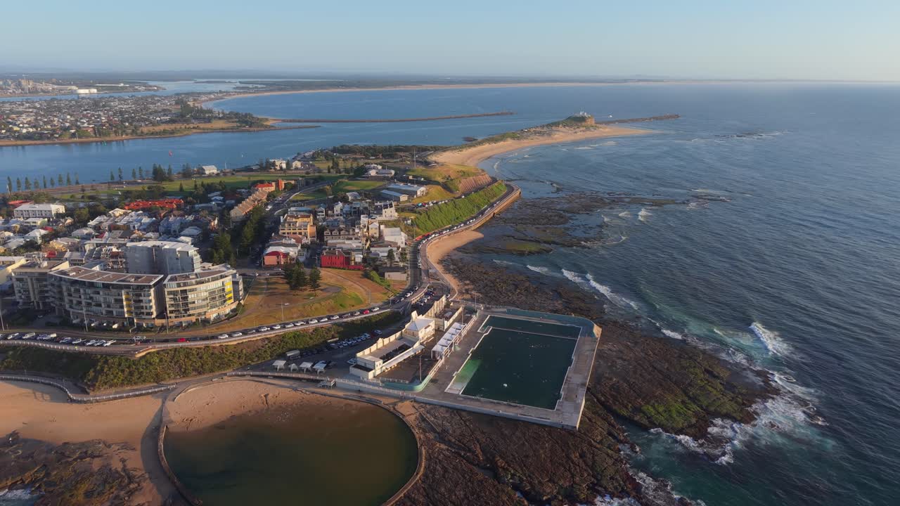 Drone pans orbit from Newcastle Ocean Baths toward Nobbys Beach on a bright sunny morning