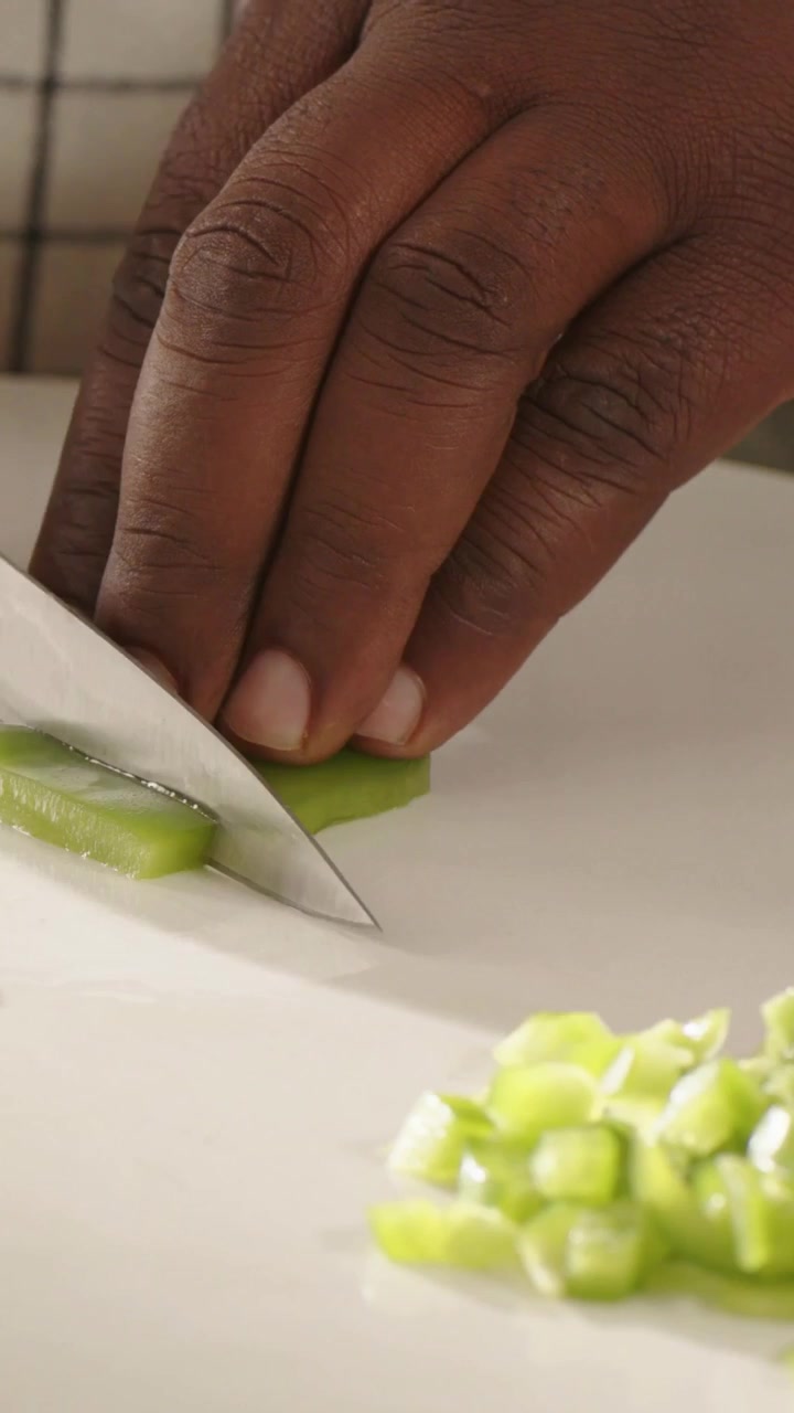 Vertikales Video: Hand kommt mit blauem Messer ins Bild, Koch würfelt grüne Paprika auf einem Schneidebrett.