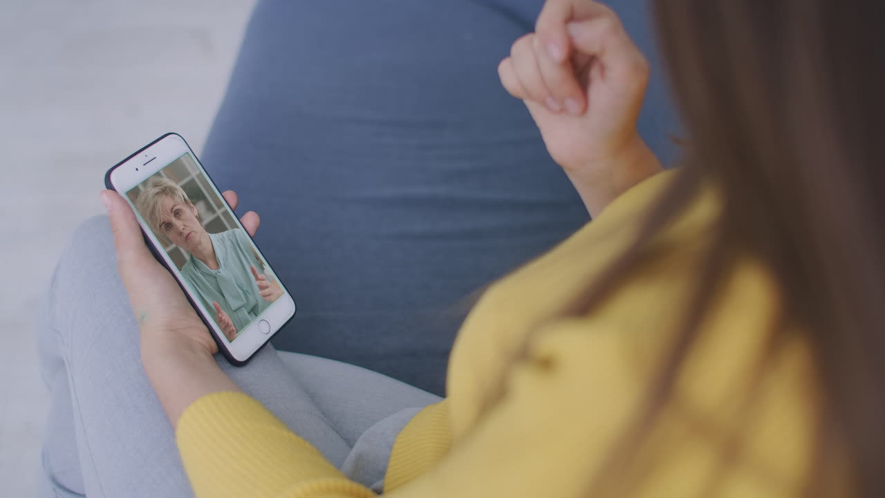 vista por encima del hombro de una mujer joven hija video llamando a padres viejos madre o amiga madura usando la aplicación de chat de conferencia en línea en la pantalla del teléfono móvil en la oficina de casa. concepto de videoconferencia familiar