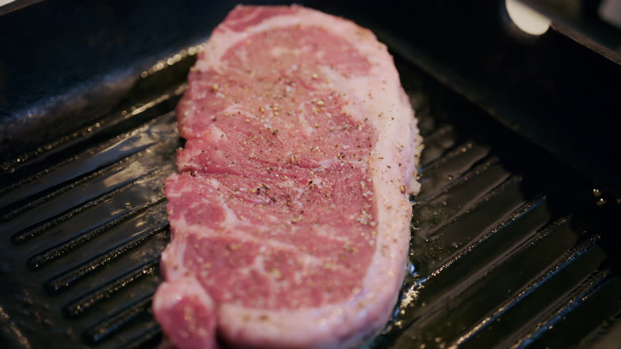 Hand placing a seasoned beef steak onto a ridged grill pan. Beginning the grilling process indoors