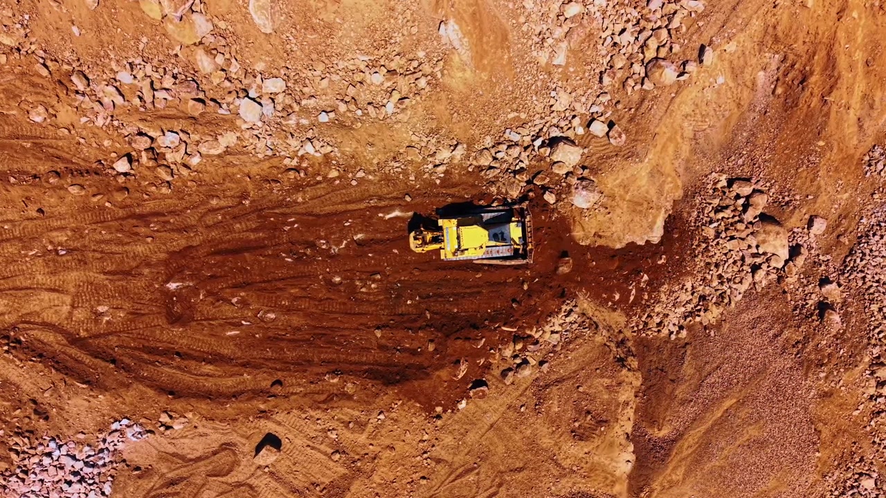 Direct overhead view of a yellow bulldozer working in dirt. A yellow crawler bulldozer clears dark soil surrounded by rocky brown ground.