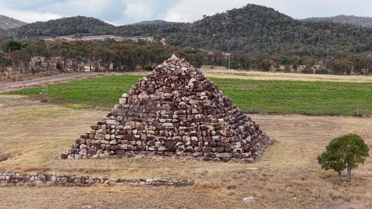 Drone camera slowly advances toward a large stone pyramid structure in an open field, surrounded by grass, trees, and distant hills under daylight