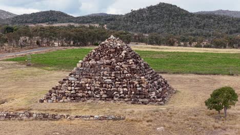 drone camera slowly advances toward a large stone pyramid structure in an open field, surrounded by grass, trees, and distant hills under daylight