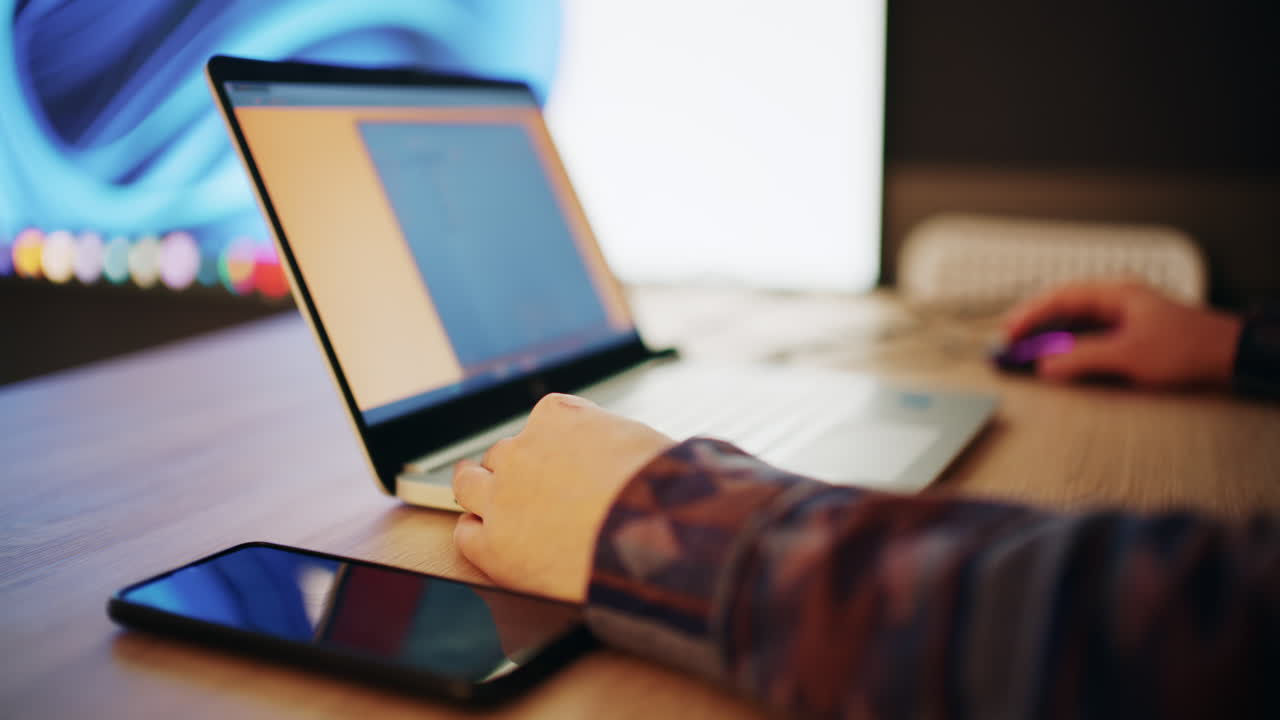 Person, der arbejder på bærbar computer ved et skrivebord med smartphone og moderne belysningsopsætning i baggrunden