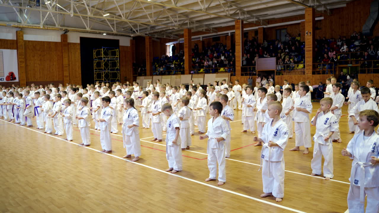 Children studying karate stand in rows in a big gym. Kids sit down on the floor together. Audience sitting at background.