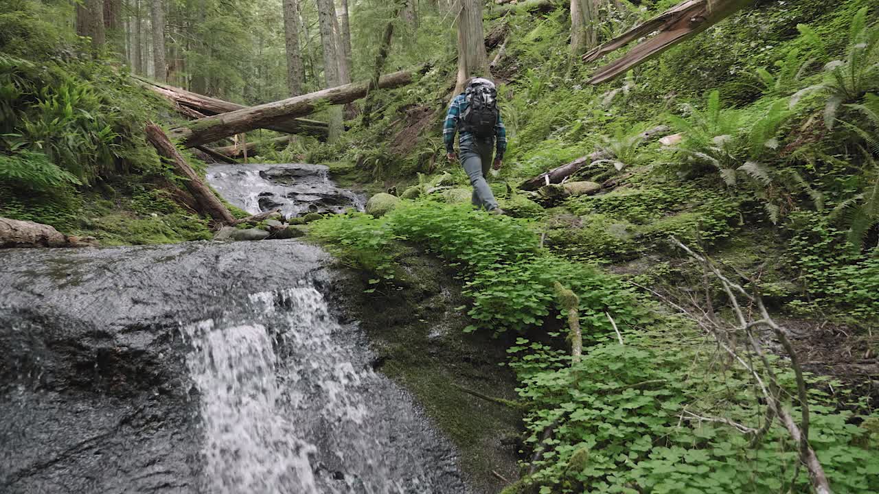 White male hiking through and up peaceful lush green forest in the distance with double waterfalls rushing stopping to enjoy the atmosphere