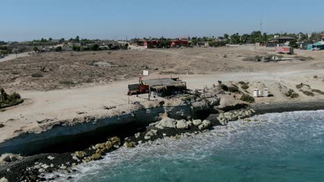 Aerial-view-Panning-shot,-Restaurant-on-the-side-of-the-rocky-Cliff-of-San-Juanico,-California-Sur,-Mexico,-rocky-shoreline-and-beach-on-a-sunny-day