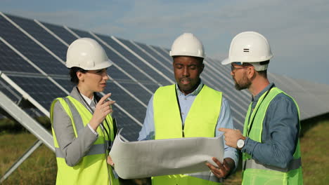 three multiethnics farm solar engineers talking while looking at blueprint on solar plantation