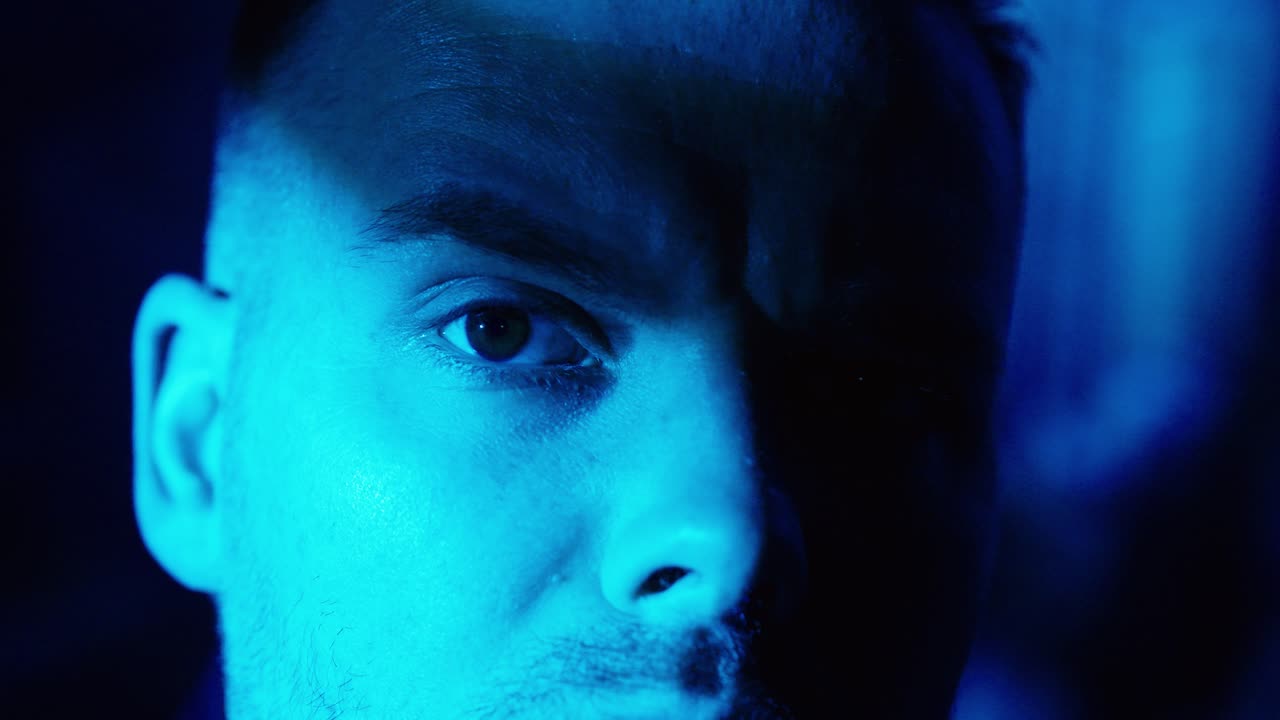 Primo piano cinematografico di un uomo che fissa intensamente la telecamera sotto un'illuminazione notturna blu, trasmettendo aggressività, minaccia, tensione e un'atmosfera da thriller psicologico o da stalker.