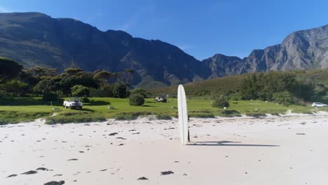 dron dando vueltas alrededor de un surfista parado en la playa mirando al agua con montañas al fondo