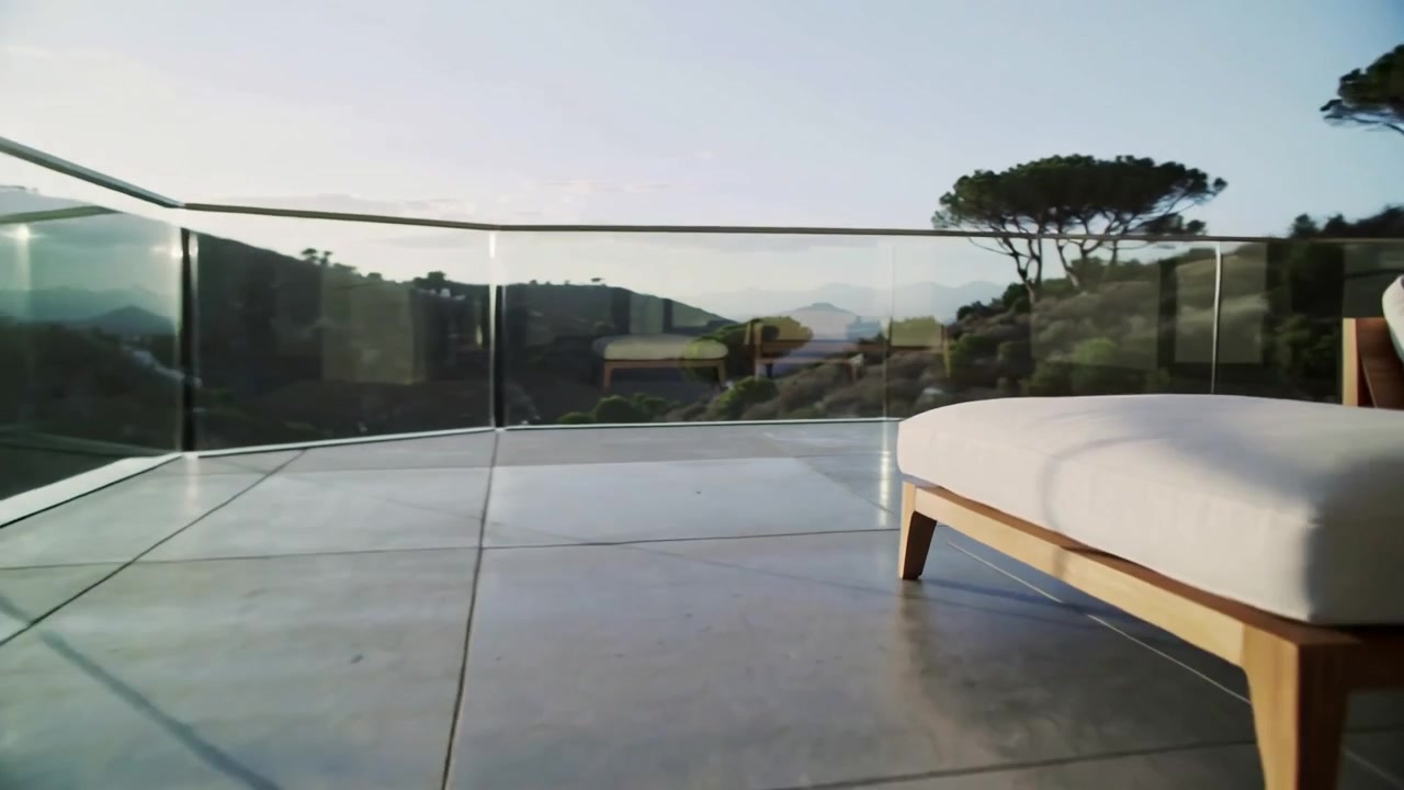 Telecamera scorrevole che si muove dall'interno verso la terrazza, rivelando una ringhiera in vetro, piastrelle in pietra e una chaise longue.