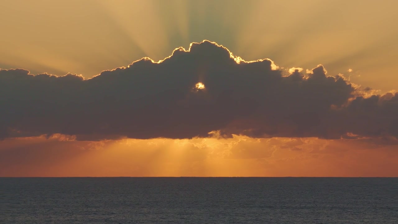Dramatischer Sonnenuntergang über dem Meer, mit goldenem Licht, das durch dunkle, silhouettierte Wolkenformationen bricht. Weitwinkelaufnahme.