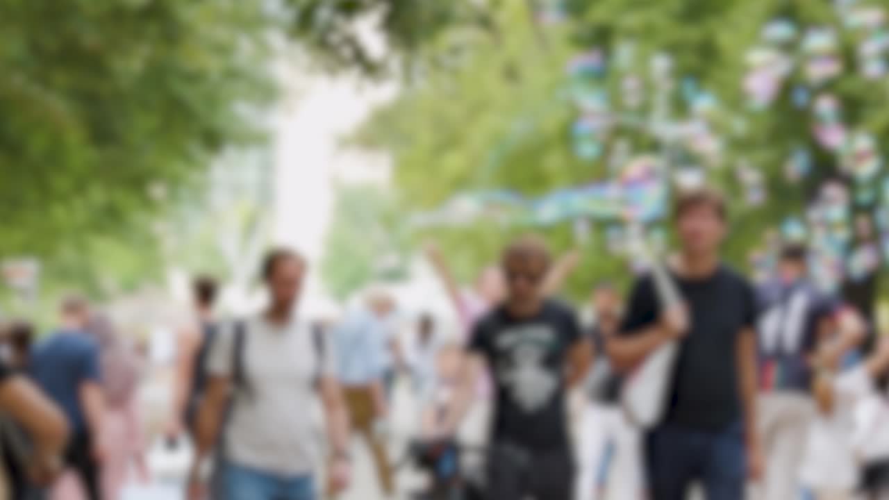 Three young adults stroll along a tree-lined park path on a sunny day, surrounded by playful soap bubbles and a lively crowd. Handheld camera, natural light