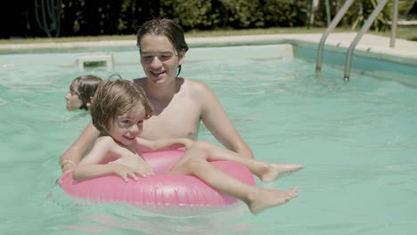 cheerful little boy floating on inflatable ring in swimming pool with the help of his brother