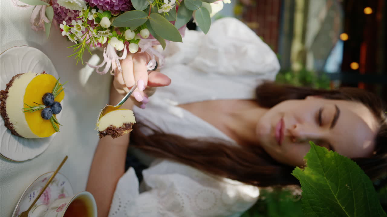 Woman eating yellow and chocolate cheese cake dessert at a terrace, composition with flowers, macaroons sweets desert and coffee and tea