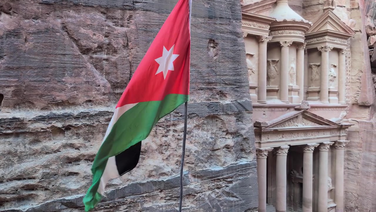 Immagini della bandiera giordana che sventola al vento con una vista dall'alto del Tesoro di Petra scavato nelle scogliere di arenaria a Petra, Giordania. Architettura antica storica e paesaggio desertico