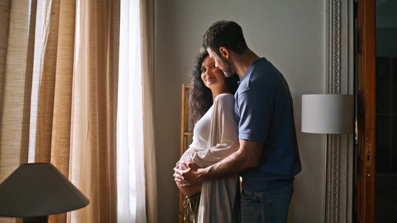 Expecting couple embracing home window. Man holds pregnant belly maternity.