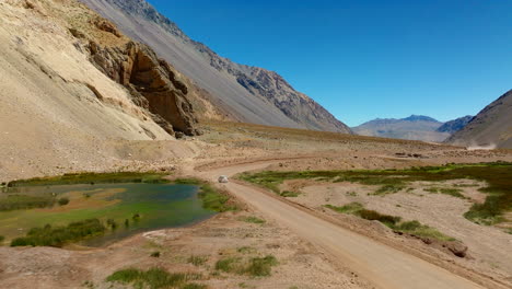 drone rearview descend to car driving along dusty road past green water through desert valley in cajon del maipo, chile