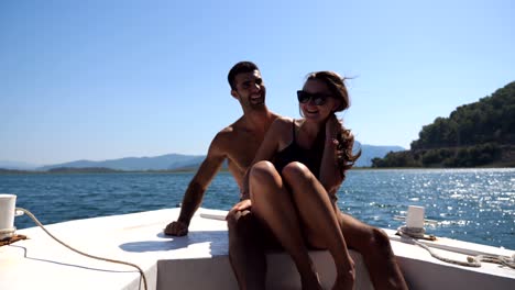 young pair sitting on bow of boat and posing to photographer at sunny day. happy couple in love spending time together on deck of ship and enjoying summer travel. concept of vacation or holiday. close up front view