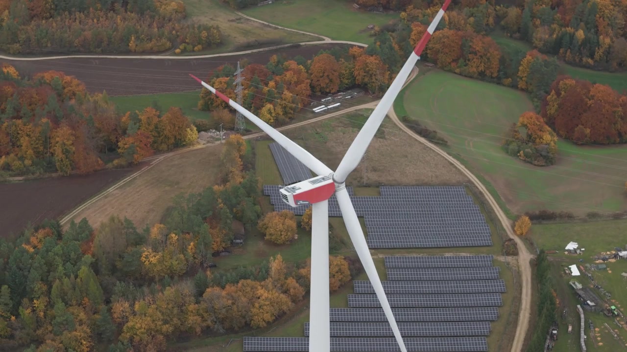 Una turbina eolica gira sopra un parco solare circondato da alberi autunnali in un paesaggio rurale.