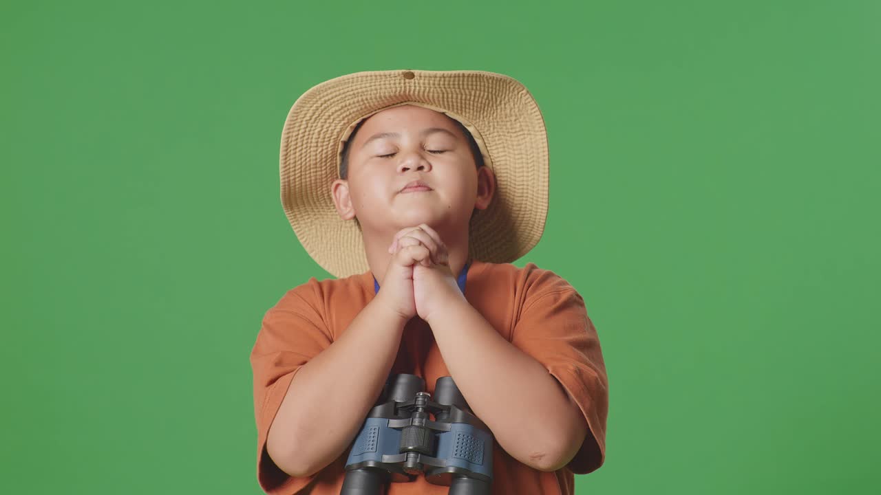 niño turista asiático con un sombrero y binoculares orando por algo mientras está de pie en el fondo de la pantalla verde. niño investigador examina algo, concepto de aventura de turismo de viaje, de cerca
