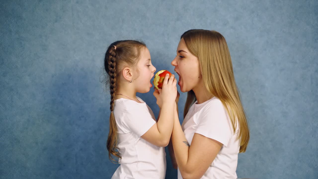 Little girl and her mother in white t-shirts bite an apple. Side view of mother and daughter eating one ripe apple on blue studio background.