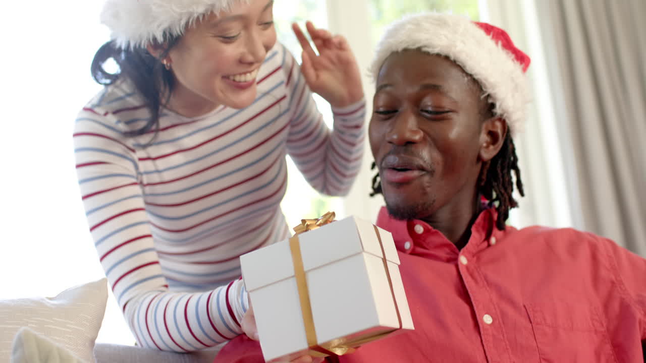 una pareja feliz y diversa con sombreros de navidad intercambiando regalos en una sala de estar soleada, en cámara lenta.