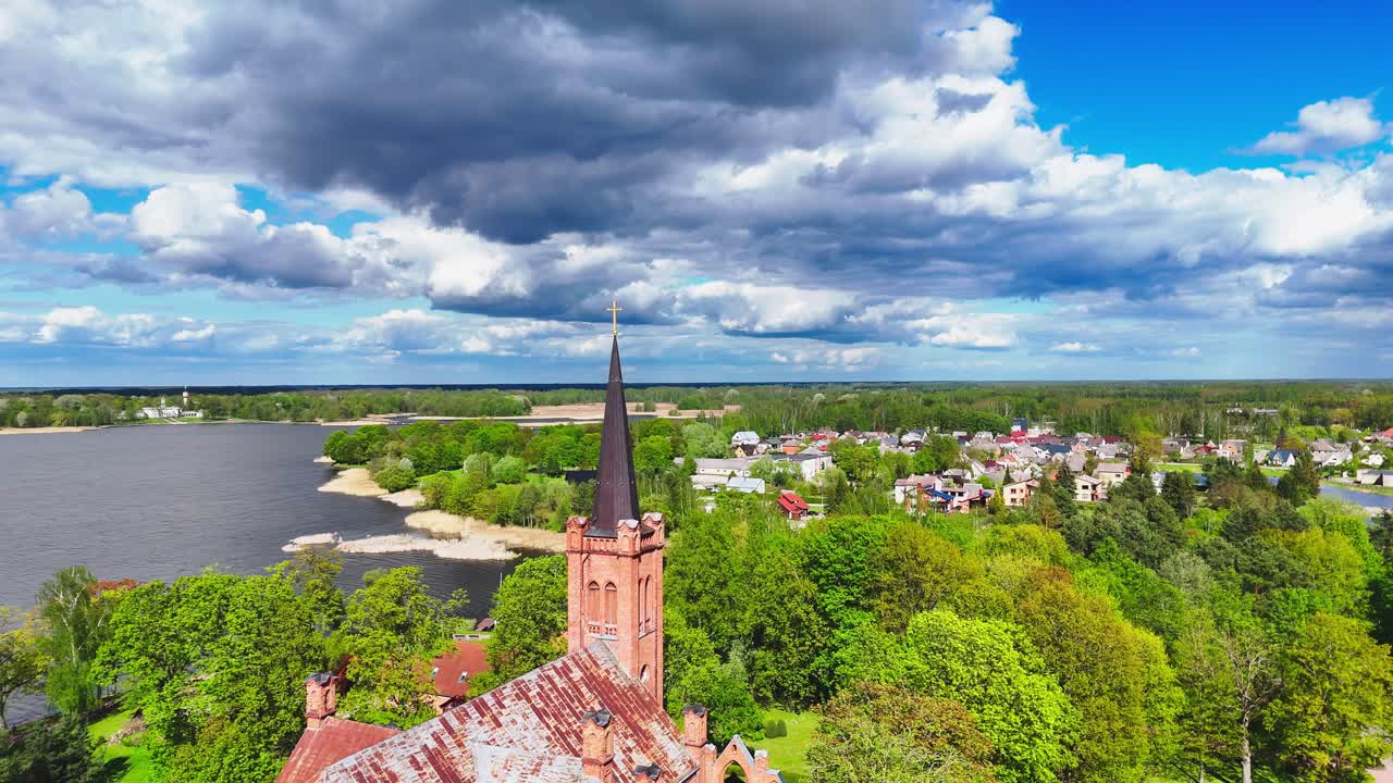 A tall church spire stands prominently above a vibrant green lakeside town, set beneath a dramatic sky filled with heavy clouds and patches of sun.