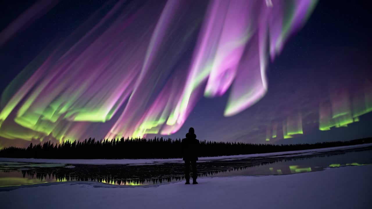 Photographer standing in snowy landscape, capturing vibrant aurora borealis reflecting on frozen lake, boreal forest silhouetted against starry night sky