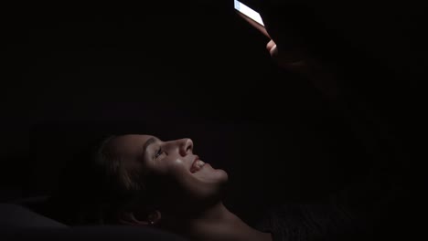 young smiling woman laying on a couch and using her phone in a dark room, chatting in the darkness with smartphone, communicating with friends online