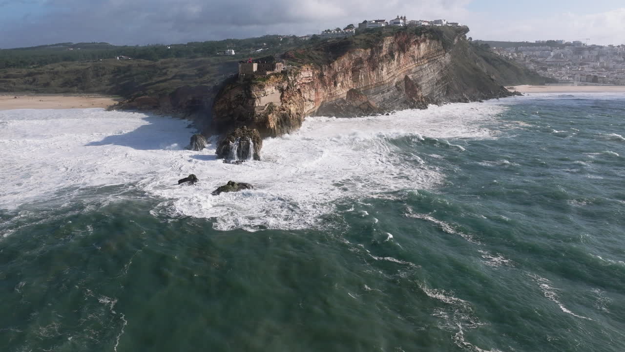 Ripresa aerea con drone di grandi onde che si infrangono su rocce e scogliere in una giornata con onde giganti a Nazaré, Portogallo, Europa. Faro di Nazaré e turbine eoliche