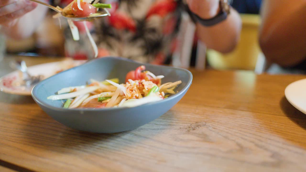 Hands serve spicy green papaya salad in a bright, casual restaurant with natural daylight