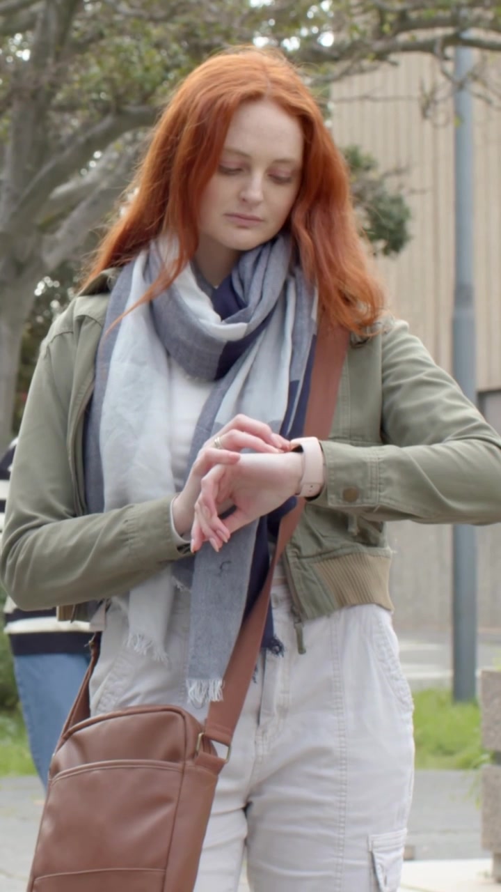 Vertical video: Approaching camera, redhead woman checking smartwatch for time while walking path