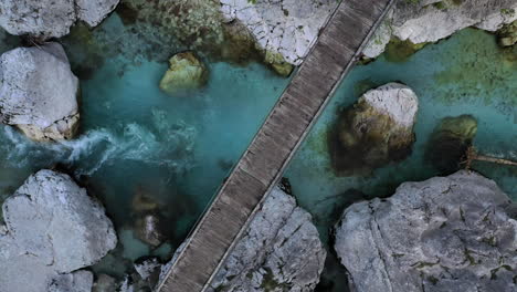 Vista-De-Arriba-Hacia-Abajo-De-Un-Puente-Peatonal-De-Madera-Que-Cruza-El-Río-Soca-En-Eslovenia-Mientras-El-Dron-Se-Eleva-Para-Revelar-El-Paisaje