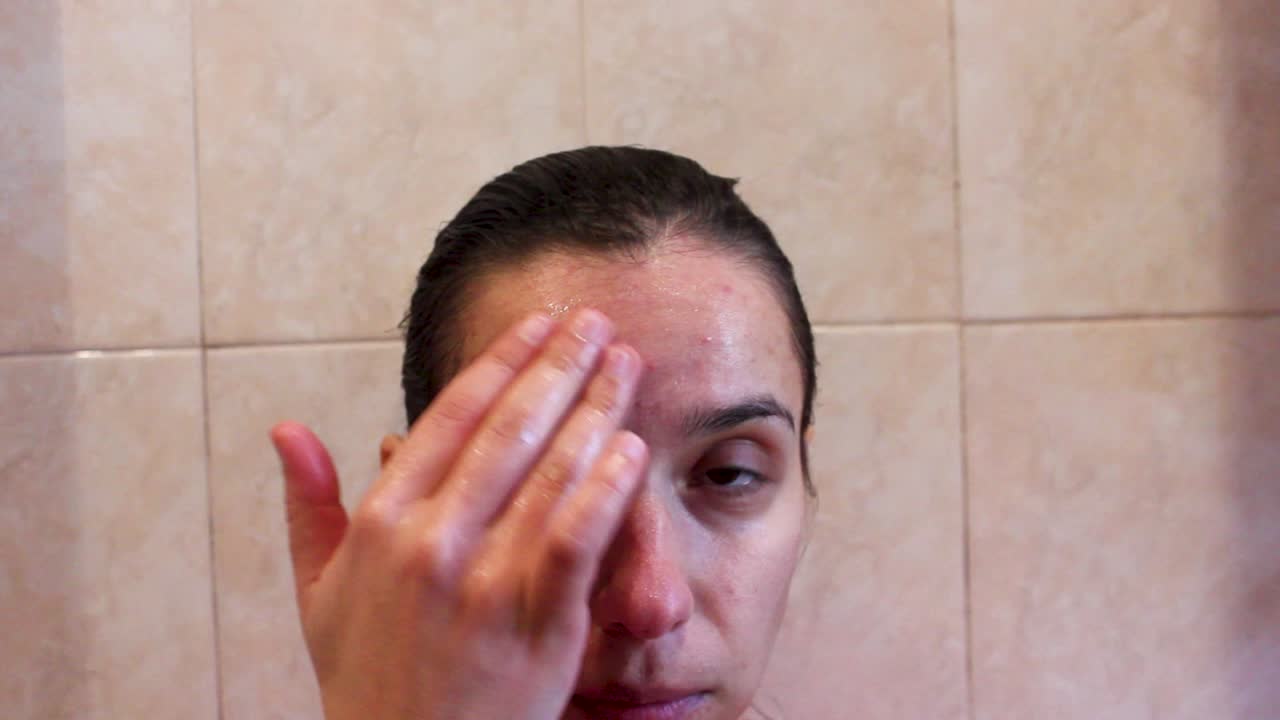mujer joven aplicando una máscara facial con las manos en la frente en el baño, de cerca