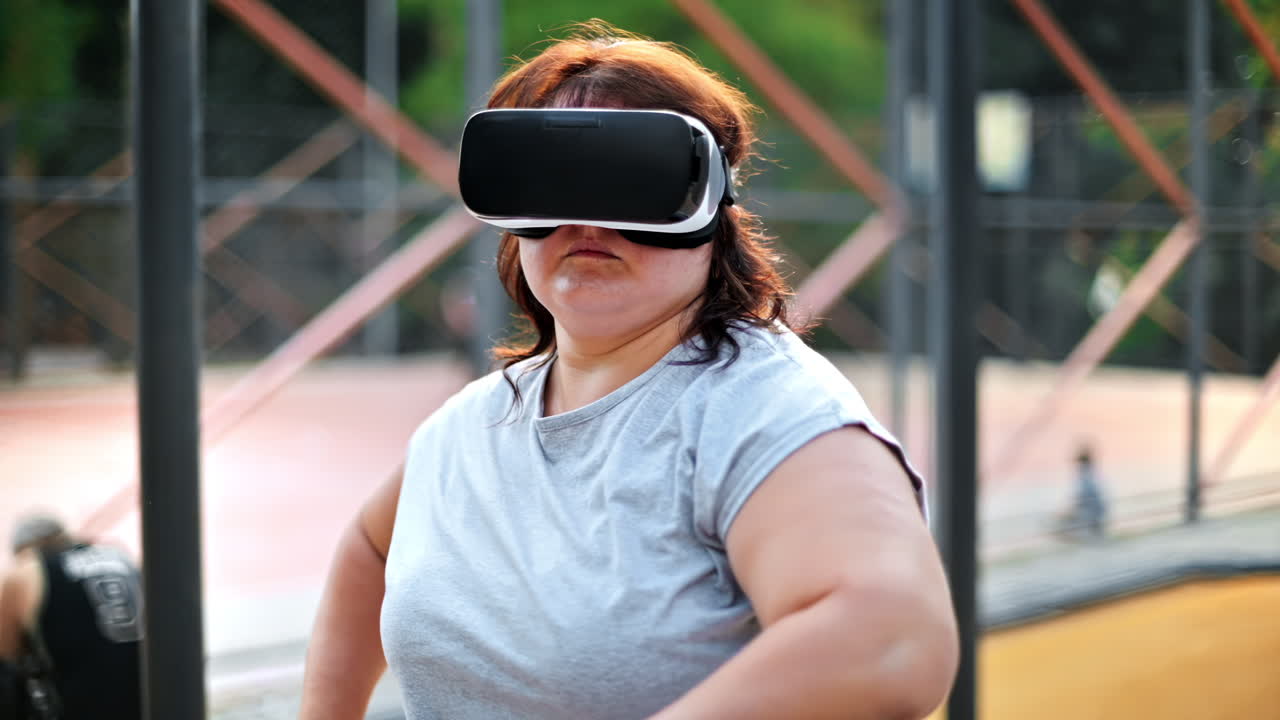 Donna in sovrappeso con maglietta grigia e leggings che si allena su un campo sportivo in un parco mentre indossa un visore per la realtà virtuale. Stile di vita fitness
