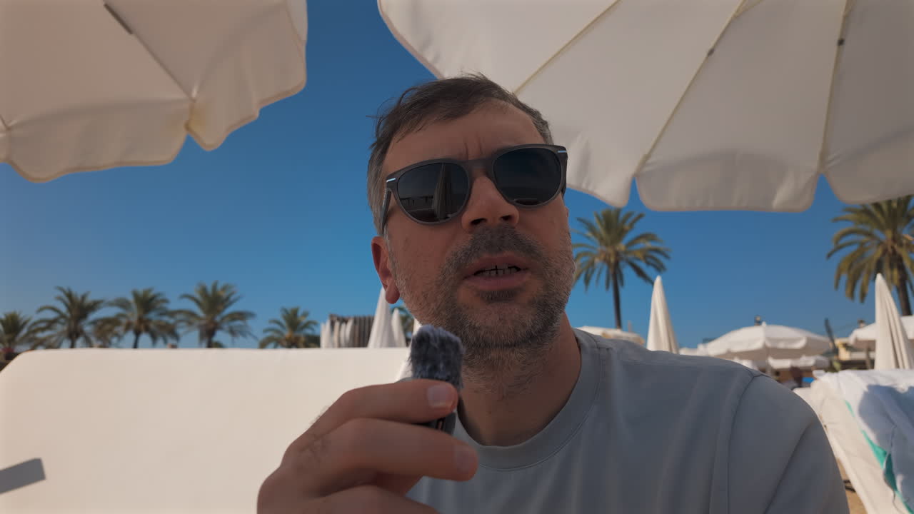 Man wearing sunglasses speaks into a small microphone under large white beach umbrellas with palm trees in the background