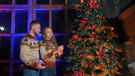 beautiful couple near the christmas tree. young woman and her husband decorating christmas tree with bright balls on large window background in the evening.