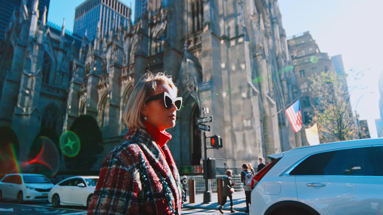 New York, USA, 5 ottobre 2025: Donna che cammina accanto all'architettura di una cattedrale gotica a New York. Donna con occhiali da sole e cappotto a quadri che cammina accanto alla Cattedrale di San Patrizio sulla Fifth Avenue a Manhattan.