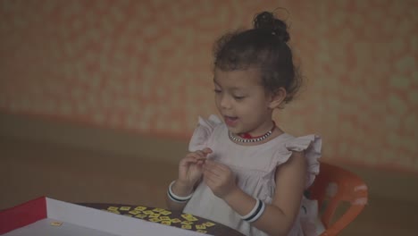 Static-handheld-shot-of-a-happy-girl-or-child-dressed-in-children's-dress-sitting-at-the-table-playing-a-game-with-letters-while-putting-words-together