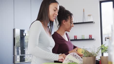 Feliz-Pareja-De-Lesbianas-Birraciales-Desempacando-Compras-Y-Hablando-En-La-Cocina,-En-Cámara-Lenta