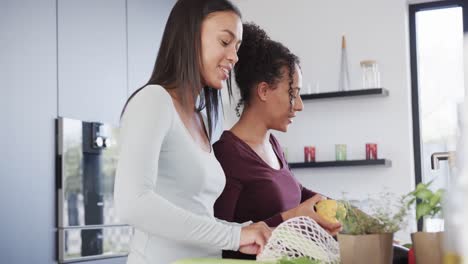 feliz pareja de lesbianas biraciales desempaquetando las compras de comestibles y hablando en la cocina, en cámara lenta