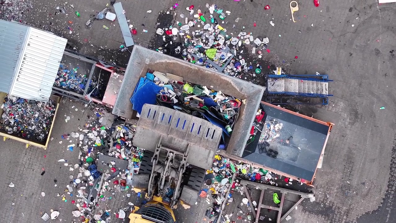 Una grande macchina elabora rifiuti plastici misti in un impianto di riciclaggio industriale. I lavoratori smistano i materiali per il recupero e il riciclaggio. L'area è trafficata da camion che consegnano i rifiuti.