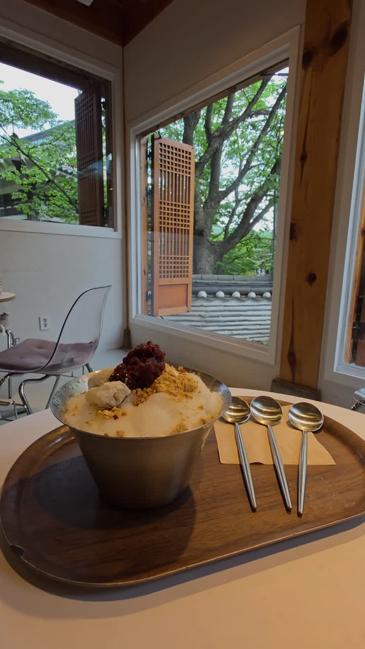 A delicious bowl of Patbingsu, a Korean shaved ice dessert with red bean and nut powder, sits on a wooden tray in a modern cafe, with a traditional Hanok village roof visible through the open window.