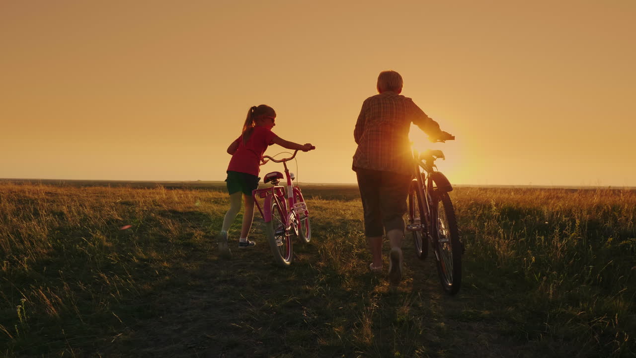 뒷모습: 한 노부인이 일몰에 손녀 자전거와 함께 걷는다 1