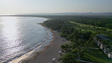 Drone-Cinematográfico-Filmado-Sobre-Playa-Dorada-Con-Reflejo-Solar-En-La-Superficie-Del-Agua-Y-El-Paisaje-Forestal-Al-Atardecer---Puerto-Plata,-República-Dominicana